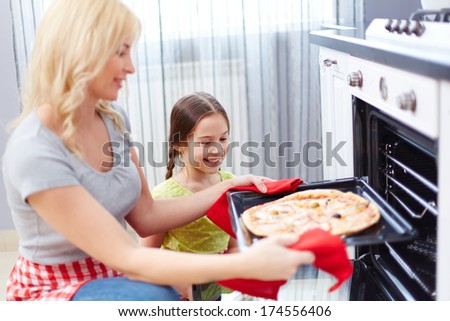Portrait of happy young woman taking pizza out of oven, her daughter standing near by