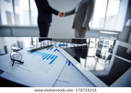 Image of eyeglasses, glass of water, touchpad and financial documents at workplace with businessmen handshaking on background