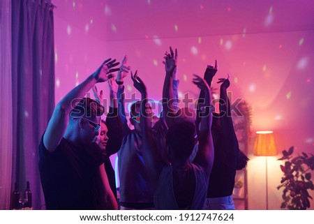 Young intercultural friends in smart casualwear raising arms while dancing together at home party in living-room illuminated with pink lighting