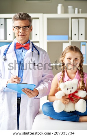 Joyful girl with teddy bear and her doctor looking at camera in hospital