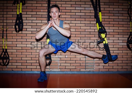 Young guy doing exercises for leg muscles over brick wall in sports gym