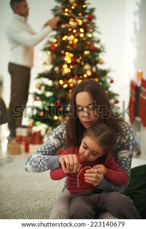 Pretty female showing her little daughter how to knit with man decorating xmas tree on background