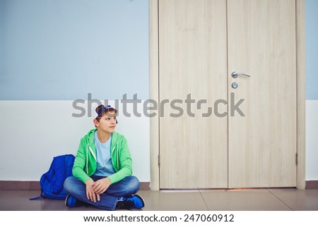 Cute schoolboy with backpack sitting on the floor by classroom door