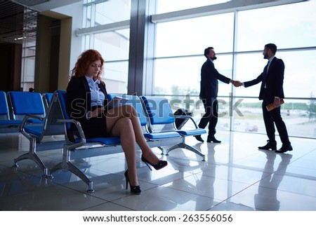 Young woman using touchpad while waiting for departure on background of two men greeting one another