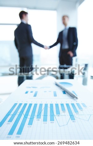 Business document with chart and graph on workplace with two men handshaking on background