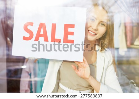 Beautiful young woman with sale announcement looking at camera from shop window