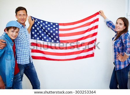 Family of three looking at camera while holding USA flag hanging on wall