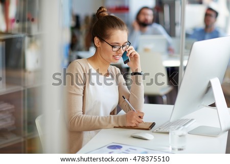 Young professional businesswoman working in public relations talking on phone with partners making notes in small notebook, sitting at computer desk in modern office space Foto d'archivio © 