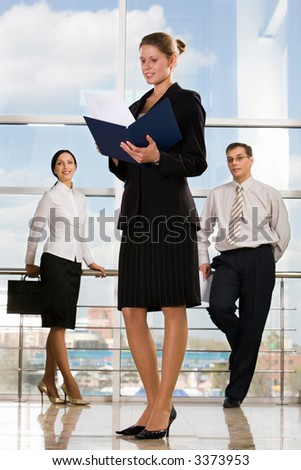Smiling Caucasian businesswoman reading documents in modern office building and two businesspeople on the background