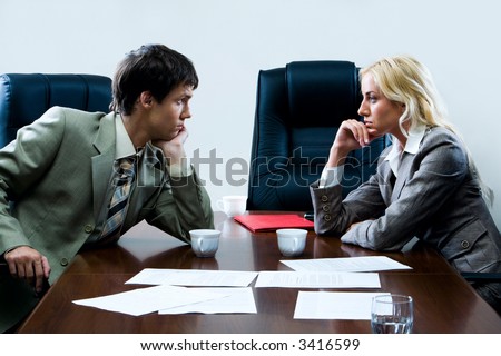 Two business people in front of each other staring hard at each other sitting at the table with cups, glass of water, paper case and littered documents on it and empty black chairs around it