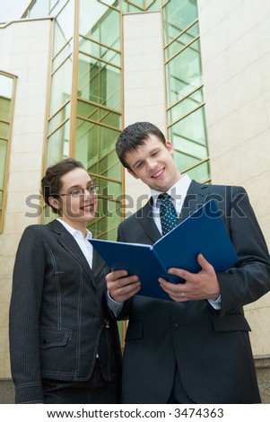Businesswoman showing documents to her colleague holding the blue paper case outside the building with glassy walls