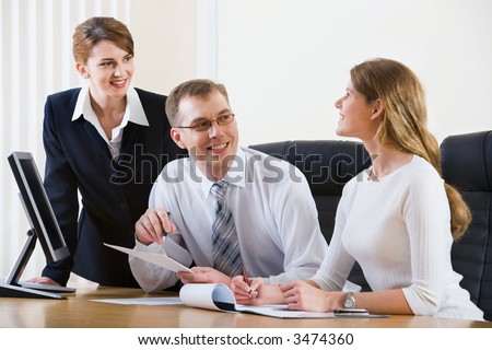 Portrait of two businesswomen and businessman discussing questions sitting at the table with a monitor and opened documents on it