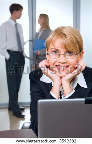 Portrait of cute smiling blond woman with eyeglasses in black suit touching her head working on laptop and two young businesspeople on the background