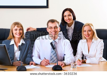 Three successful smiling people sitting in a row in  chairs at the table with monitor, microphones and documents on it and cute business woman standing behind them looking at the camera