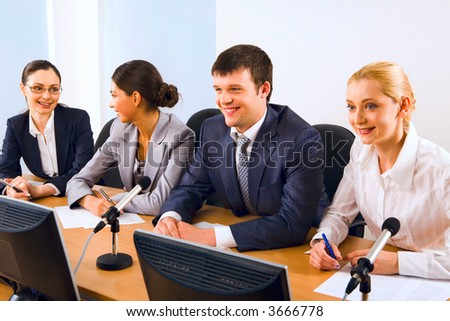 Four successful young people sitting in a line in front of monitors and microphones speaking to each other sitting at the table in black comfortable chairs