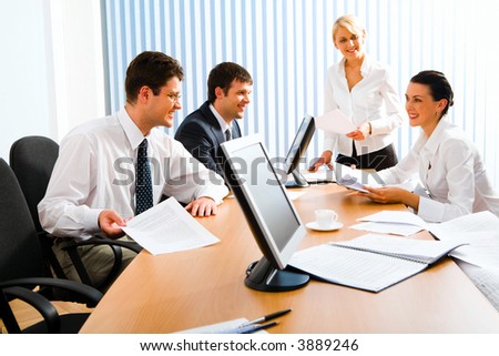 Team of successful business people sitting at the table with monitors, notepad, papers and white cup on it talking to each other discussing important questions
