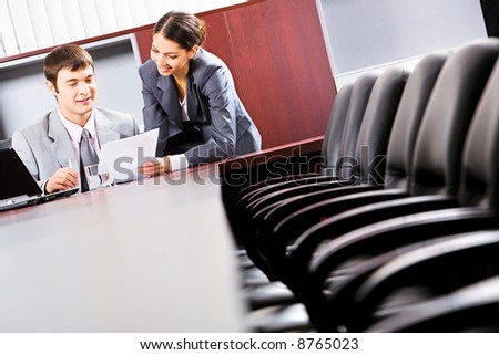 Image of a business man sitting and reading a document in the office with a smiling business lady standing near by