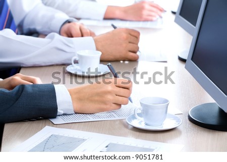 Hands of three business people over the documents lying on the table with cups and monitors near by