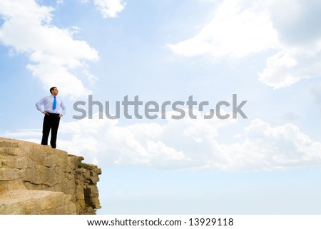 Image of successful businessman standing on rock and looking straight on the background of sky