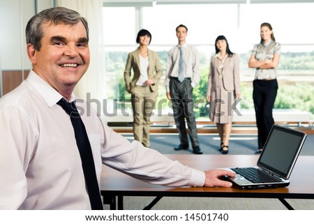 Photo of older smiling leader touching the laptop on the background of working team