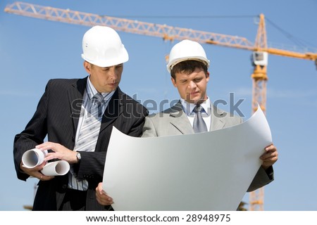 Portrait of two confident foremen looking at project on the background of sky and crane