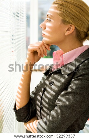 Profile of serious woman looking through venetian blind and thinking about something