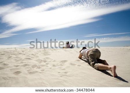 Careful photographer taking shots of relaxing couple sitting back to back on sandy beach against blue sky