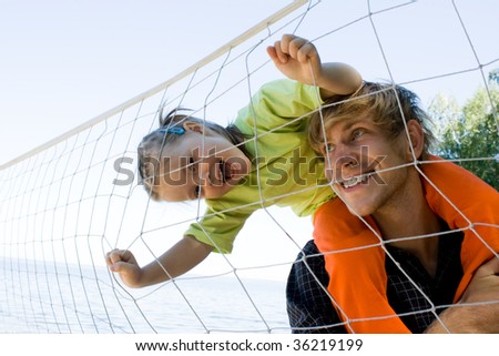 Little girl laughing on young man neck and looking at camera through volleyball net