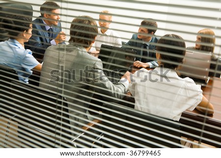 View from behind venetian blind of associates interacting at working meeting