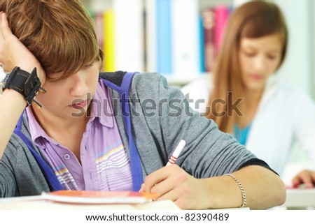 Portrait of smart guy making notes in copybook with his classmate behind