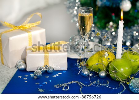 Image of holiday table with flute of champagne, fruits, gifts, burning candle and decorations on it