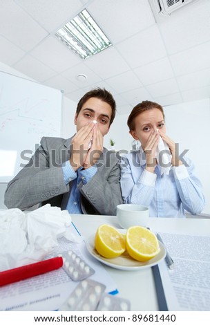 Image of sick business partners blowing their noses and looking at camera in office
