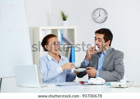 Image of businessman sneezing while his anxious partner giving him a cup of tea in office