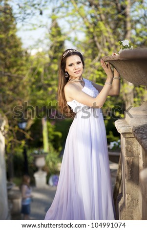 beautiful slim bride in luxury dress in park on sunset near blossom flowers in wedding day. young woman in Greek goddess style with diamond tiara and jewelery outdoor. romantic girl with glossy hair.