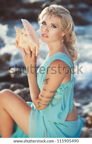 Alluring blonde woman with big seashell in Greek Goddess style at sunset. Beautiful girl in blue silk luxury dress with golden jewelry. freedom and romantic concept. positively thinking, Sea Nymph.