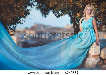 alluring blond woman outdoor in Greek goddess style with golden jewelery in luxury dress on sunset near ancient ruins. stylish romantic girl in blue silk. romantic concept. Amazing Nymph. Greece
