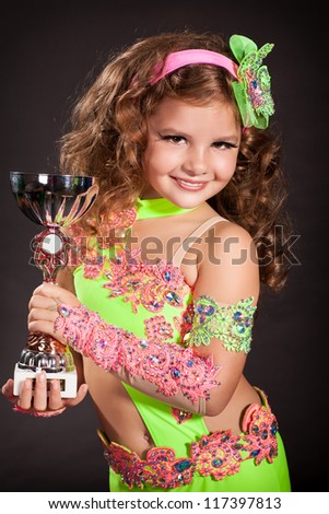 Adorable toddler girl bellydancer posing with cup in traditional oriental costume. Beautiful arabian little child with curly hair. Portrait of baby artist in carnival latin dress. Girl contest winner.