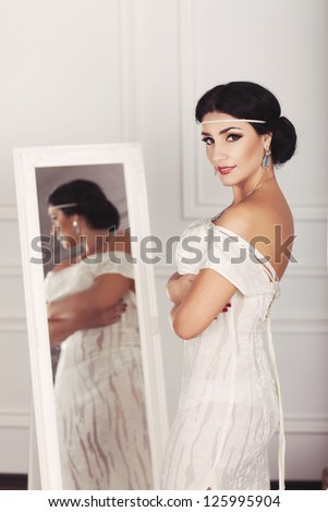 Gorgeous bride with bright makeup and fashion greek hairstyle in white wedding dress and jewelry waiting for groom at home. Romantic stylish woman in vintage dress have final preparation for wedding