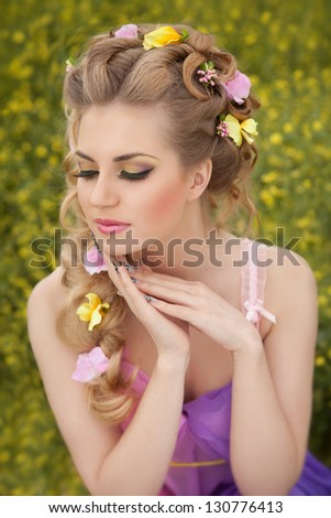 Pretty blond woman at spring blossom field with flowers in hair Bright make-up outdoors. Alluring girl with gorgeous fantasy hairstyle posing at summer nature. Sexy romantic young lady. Spring concept