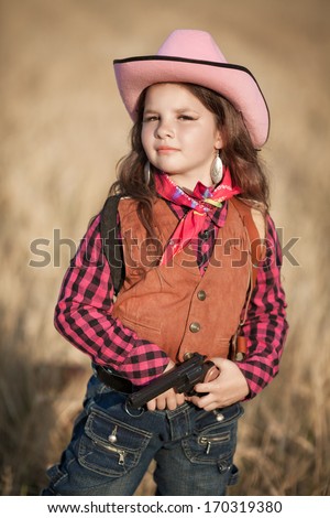 Cute Child having fun outdoors, little girl cowboy playing in wheat field at sunset. Happy baby girl with toy gun and cowboy hat enjoying nature. American Cowgirl. lovely smiling toddler portrait.