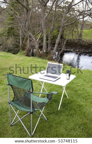 table chair and laptop computer outside on the grass by a river