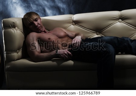Muscular topless male bodybuilder laying on a leather sofa.