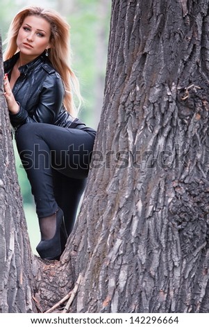 Stylish strawberry blonde woman or model in black leather and heels leaning on a tree trunk.