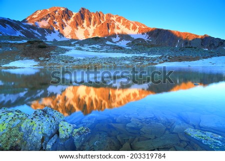 Orange light on distant summit reflected by calm mountain lake