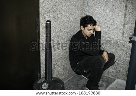 A young asian guy is squatting in a corner, his arm supporting his head and thinking/Portrait of a young asian guy
