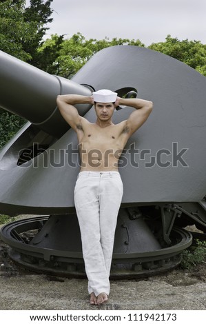 A masculine guy, wearing a marine hat and half naked, is leaning on a big gun, hands on the back of his head and thinking.../On the military base