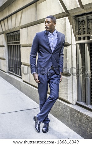 A young black businessman is standing outside by a window in a street and taking a break/Portrait of Young Black Businessman