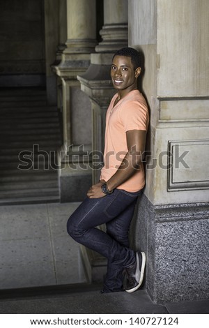 Leaning on a column, a young handsome black guy is standing at a hallway and smilingly looking at you./Portrait of Young Black Guy