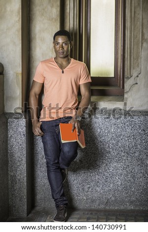Standing by a window and holding a red book, a young handsome black guy is reading and relaxing outside/Read Outside