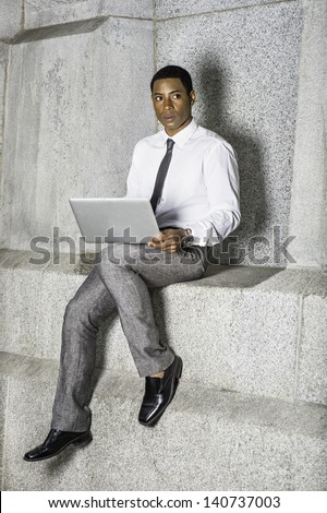 Dressing in white shirt, a black tie, gray pants, a young handsome black businessman is sitting outside and working on a computer./Work Outside